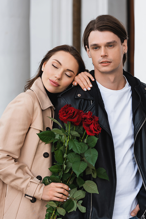 pretty woman in trench coat holding red roses and leaning on shoulder of boyfriend in stylish jacket