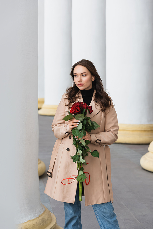 pretty young woman in beige trench coat holding red roses and looking away