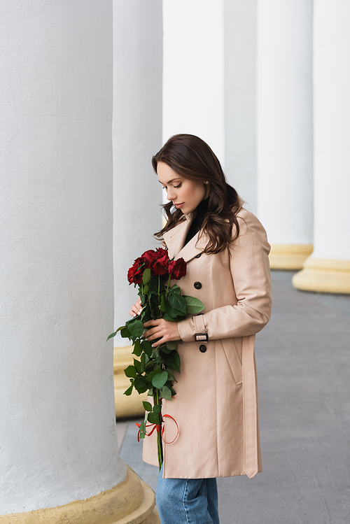 pretty young woman in beige trench coat looking at blooming red roses