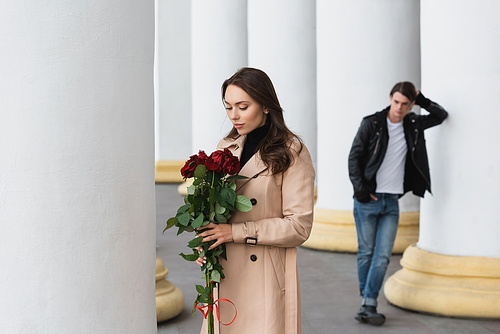 pretty young woman in beige trench coat looking at red roses near boyfriend on blurred background