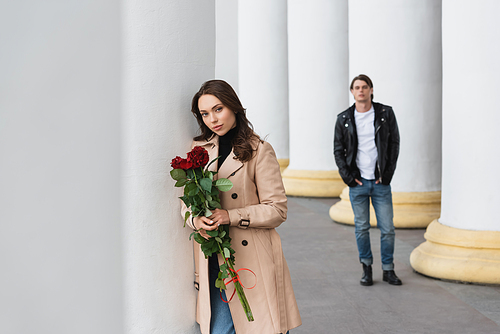 pretty woman in beige trench coat holding red roses near boyfriend on blurred background