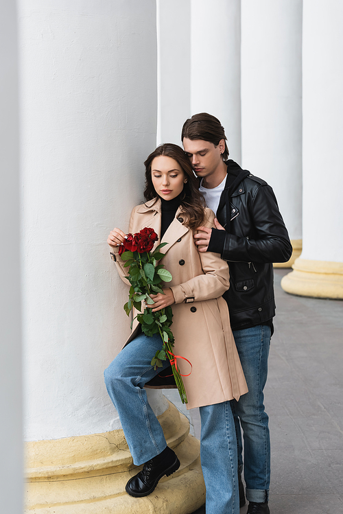 full length of stylish young man hugging pretty woman in beige trench coat holding red roses