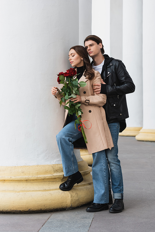 full length of stylish man hugging pretty girlfriend in trench coat holding red roses