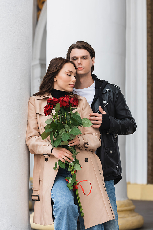 stylish man hugging pretty girlfriend in stylish trench coat holding red roses