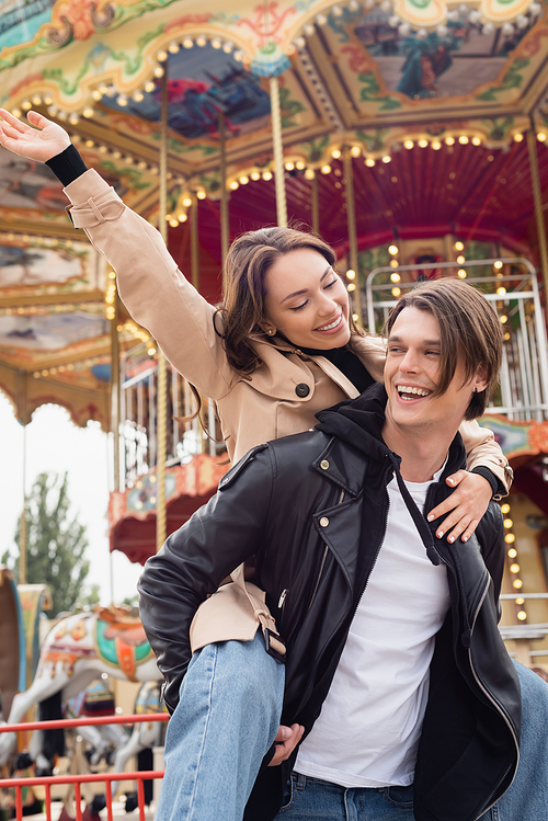 stylish man in black jacket piggybacking happy girlfriend in amusement park