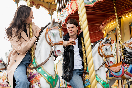 cheerful young couple in stylish outfits holding hands and riding carousel horses in amusement park