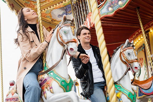 happy young couple holding hands and riding carousel horses in amusement park