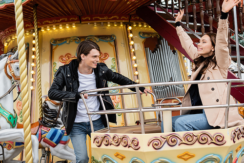 cheerful man in trendy outfit looking at happy girlfriend with raised hands on carousel in amusement park