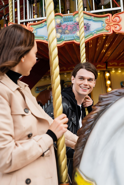 happy young man looking at girlfriend riding carousel horse in amusement park