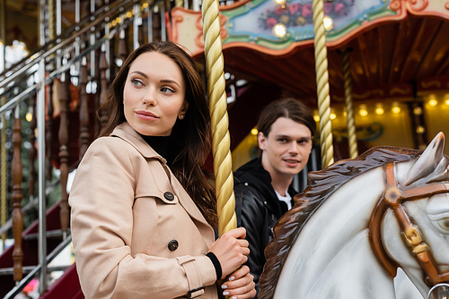 pretty woman in trench coat riding carousel horse near blurred boyfriend in amusement park