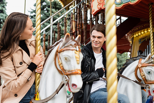 cheerful young man looking at girlfriend riding carousel horse in amusement park