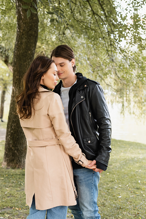 pretty young woman in trench coat and cheerful man in jacket holding hands in park