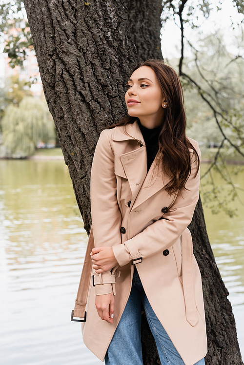 pleased young woman in beige trench coat leaning on tree trunk in park