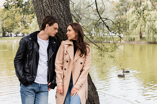 stylish man and pretty woman in trench coat looking at each other near river in park