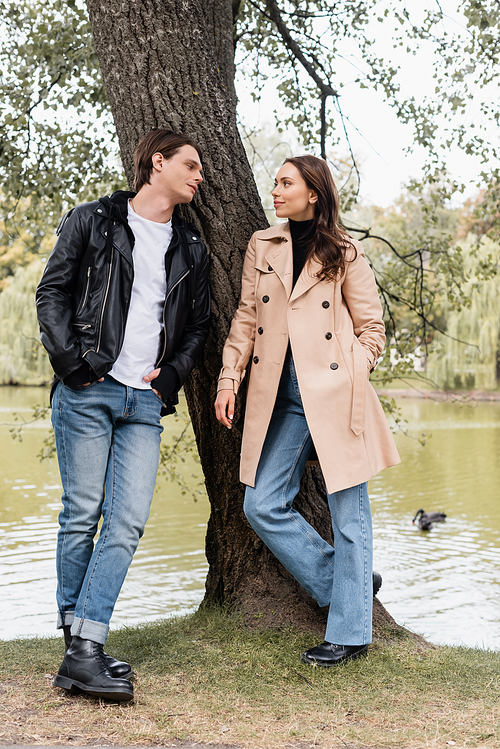 full length of stylish man and pretty woman in trench coat looking at each other near river in park