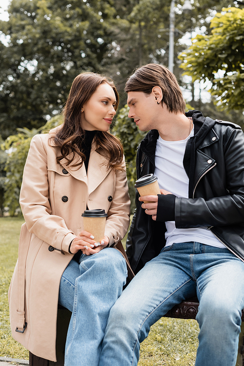 young and stylish couple holding paper cups with takeaway drink while sitting on bench in park