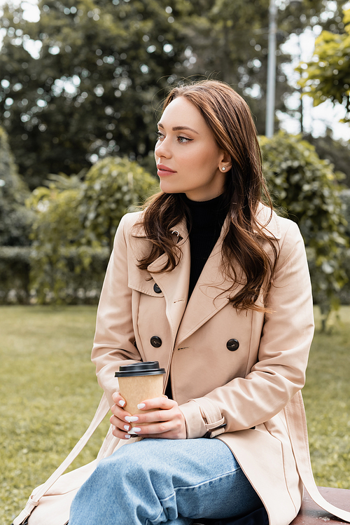 pretty young woman in beige trench coat sitting on bench with paper cup