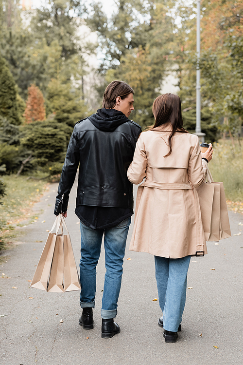 back view of woman in trench coat holding paper cup near boyfriend with shopping bags while walking in park