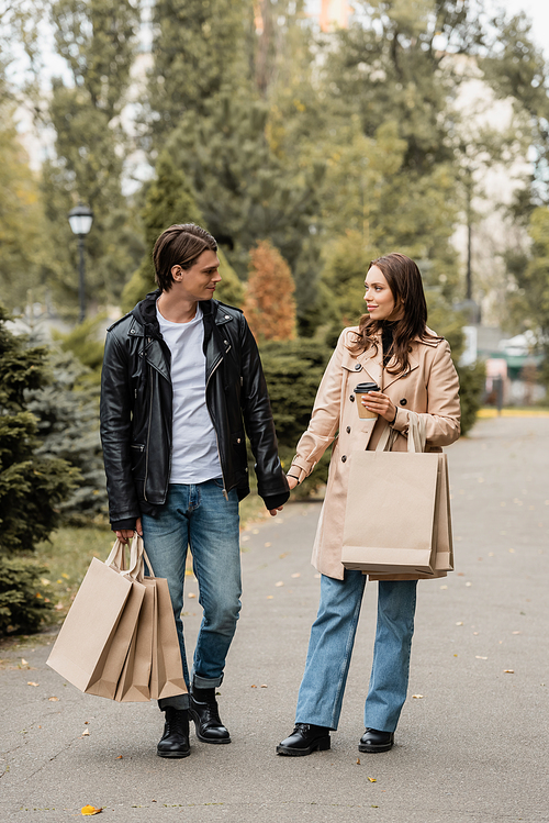 full length view of woman holding paper cup and hand of boyfriend with shopping bags  in park