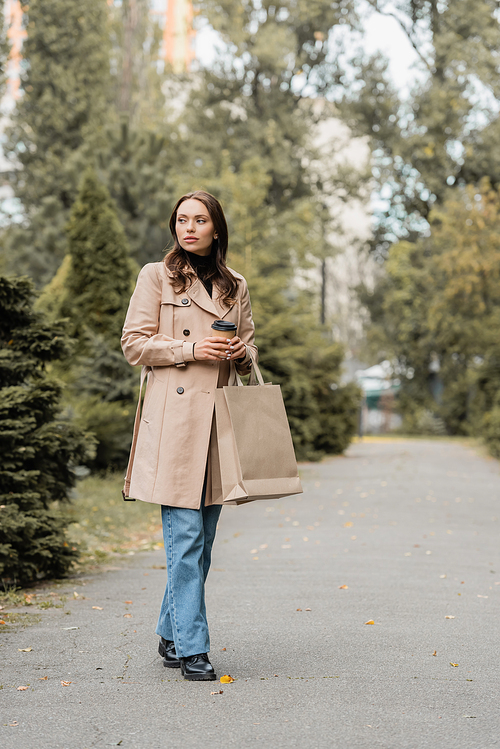 full length view of young woman in trench coat holding paper cup and shopping bags  in park