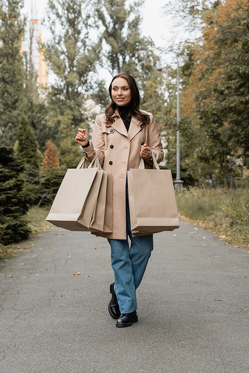 full length of happy young woman in trench coat holding shopping bags in autumnal park