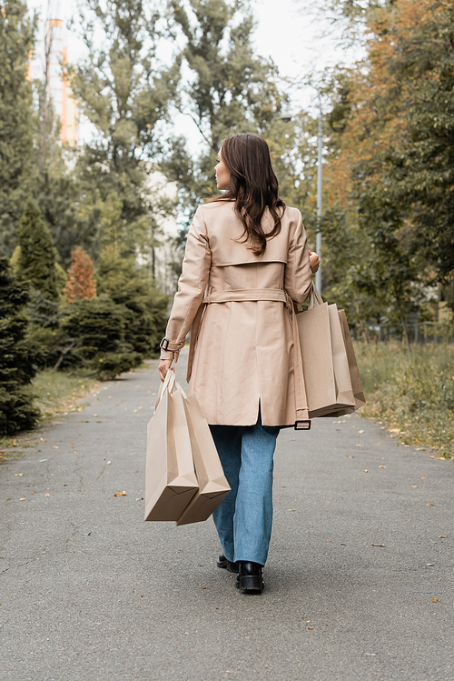 full length of young woman in trench coat holding shopping bags and walking in autumnal park