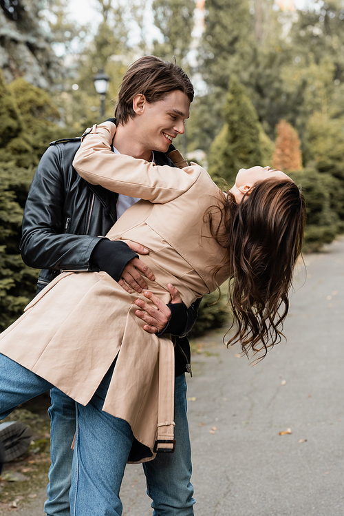 cheerful man dancing with brunette girlfriend in trench coat in autumnal park