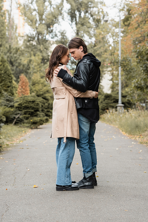 full length view of stylish young man hugging pretty girlfriend in trench coat outdoors