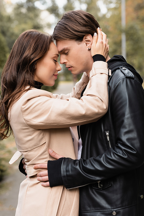 side view of pleased woman in beige trench coat hugging boyfriend in black jacket