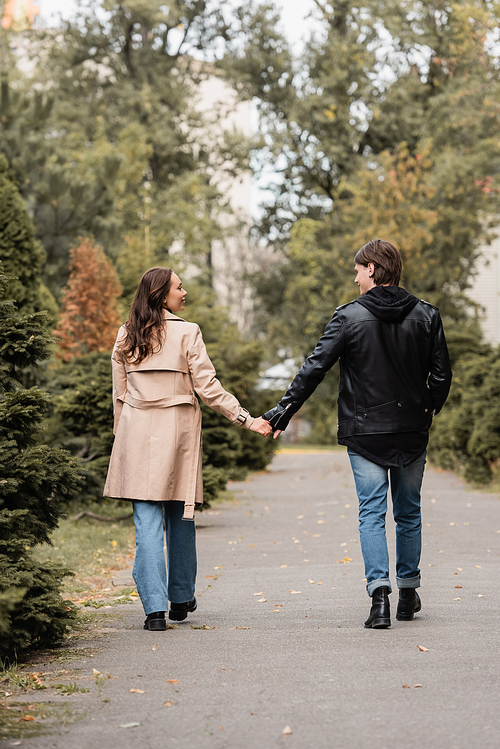 full length of young and stylish couple in autumnal outfits holding hands while walking in park