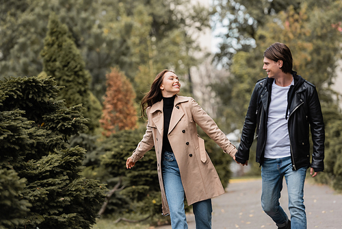 cheerful and stylish couple in autumnal outfits holding hands while walking in park