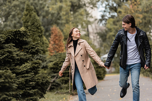 joyful and stylish couple in autumnal outfits holding hands while walking in park