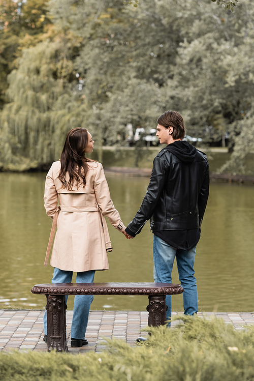 back view of young couple in stylish outfits holding hands while standing near lake in autumnal park