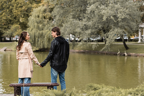 young couple in autumnal outfits holding hands while standing near lake in park