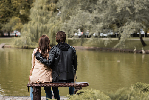 back view of young man in black jacket hugging girlfriend while sitting on bench near lake in park