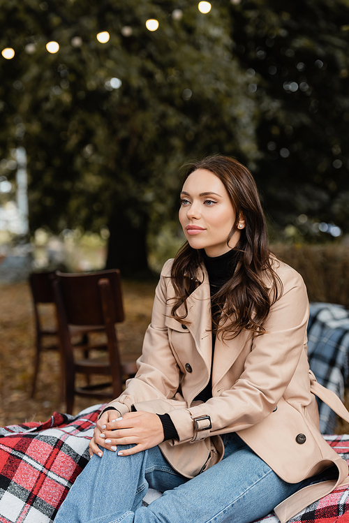 dreamy young woman in beige trench coat sitting on blanket during picnic in park