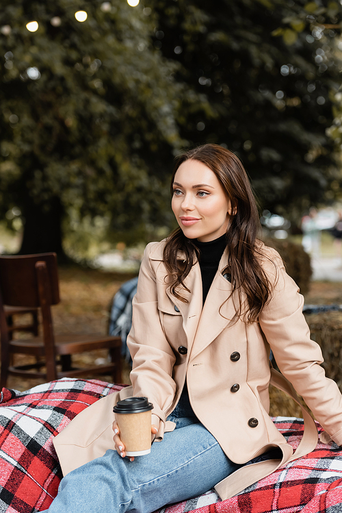 cheerful young woman in beige trench coat sitting on blanket and holding paper cup