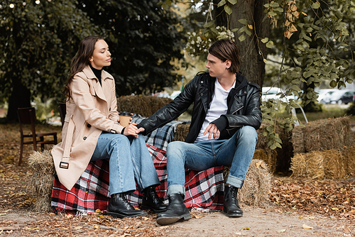 stylish man holding hand of girlfriend sitting with paper cup on plaid blanket