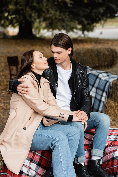 stylish man in black jacket hugging girlfriend in trench coat while sitting on blanket in park