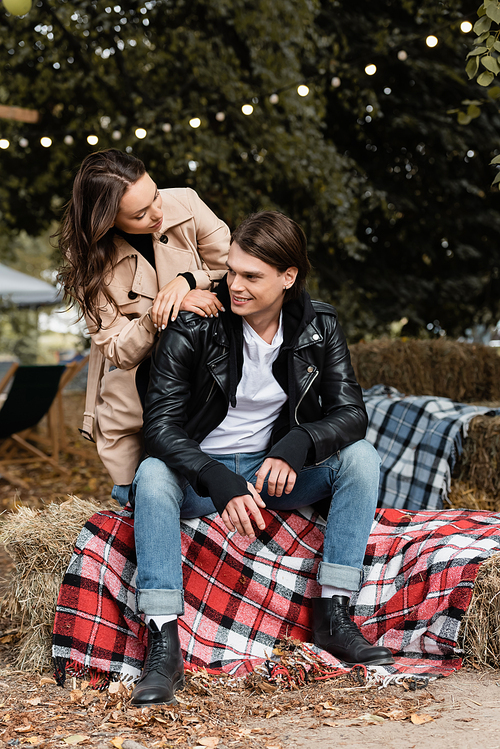 young woman in trench coat looking at cheerful man sitting on blanket in park