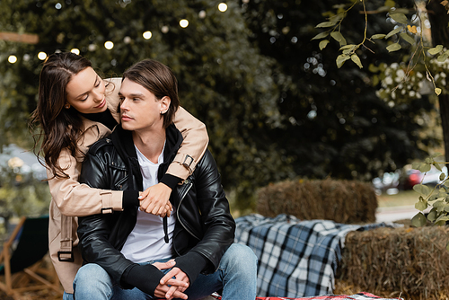 young woman in trench coat hugging stylish boyfriend sitting with clenched hands in park