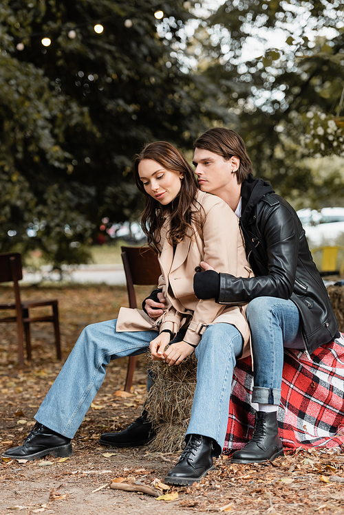 stylish man in black jacket hugging young girlfriend in trench coat sitting in park