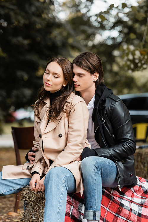 stylish man in black jacket hugging young girlfriend with closed eyes sitting in park