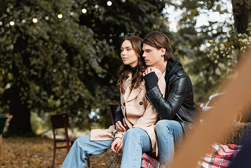 stylish man in jacket hugging young girlfriend in beige coat sitting in park