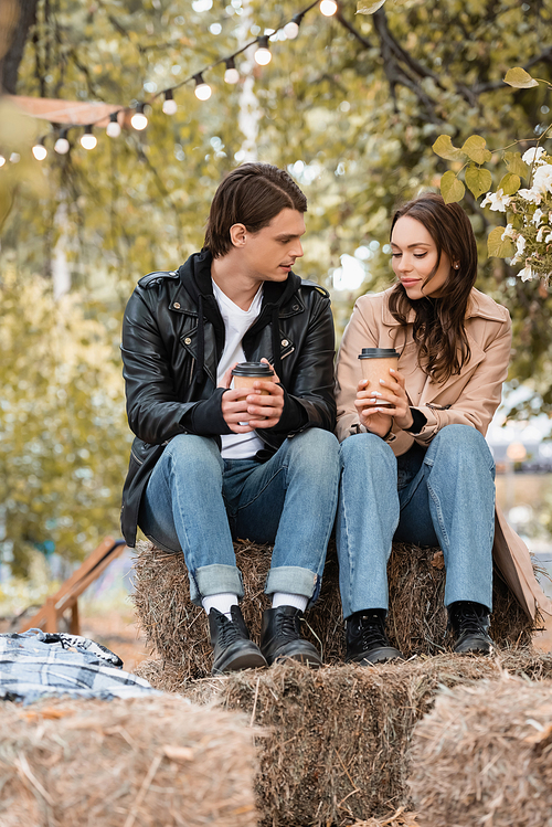 young woman and man in stylish outfits holding paper cups and sitting in autumnal park