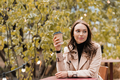 dreamy woman in beige trench coat holding paper cup with coffee to go in autumnal park