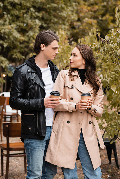 young woman and man in stylish outfits holding coffee to go and looking at each other