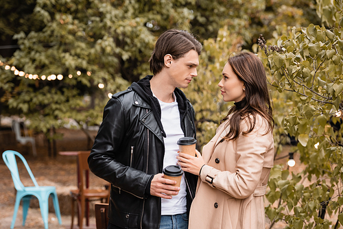 side view of young woman and man in stylish outfits holding coffee to go and looking at each other in park