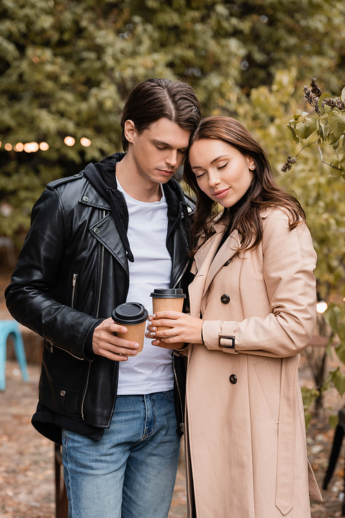 young woman and man in stylish outfits standing with closed eyes and holding coffee to go