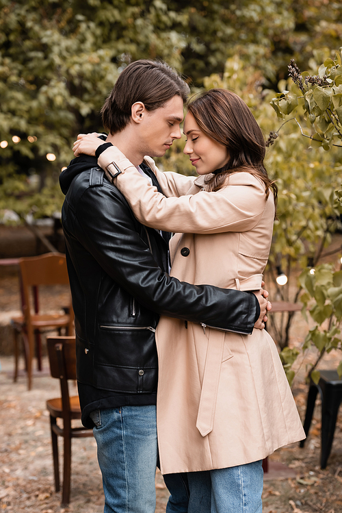 side view of couple with closed eyes standing in stylish outfits and hugging in autumnal park